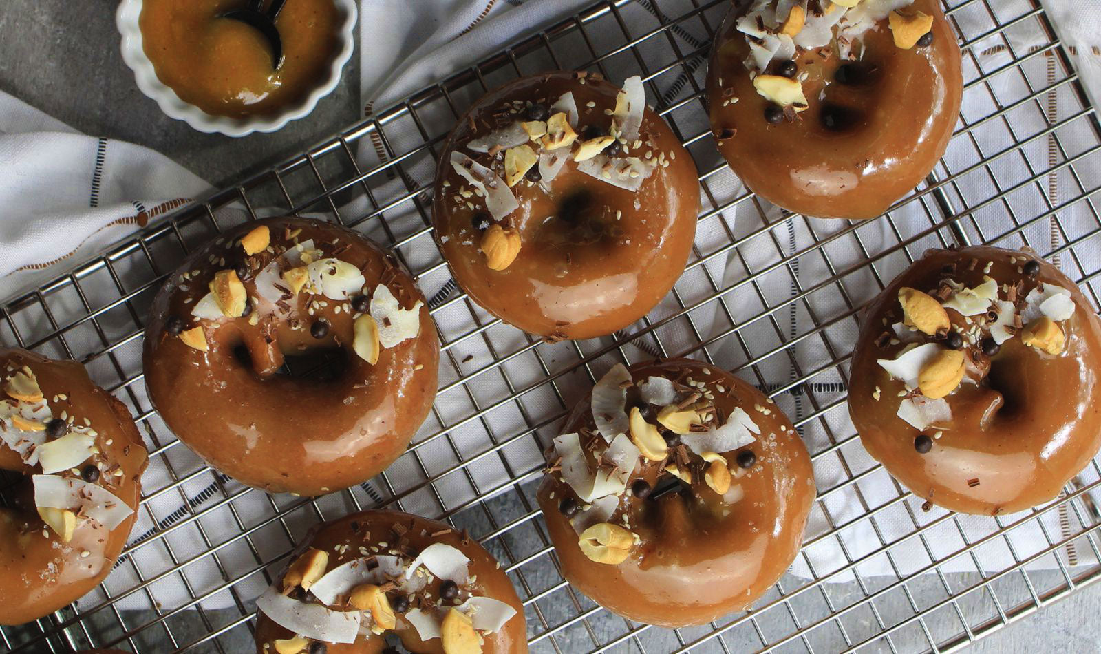 Maple cashew glazed donuts on a cooling rack decorated with cashews, coconut, chocolate and sprinkle of sea salt.