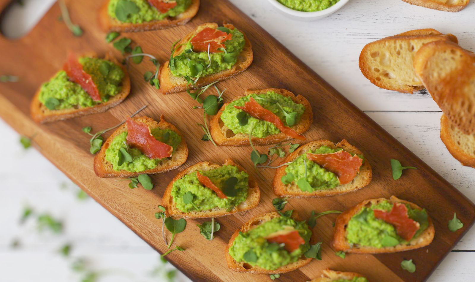 Vibrant green Pea Pesto atop a lightly toasted crostini and garnished with crispy prosciutto on a warm wooden serving platter.