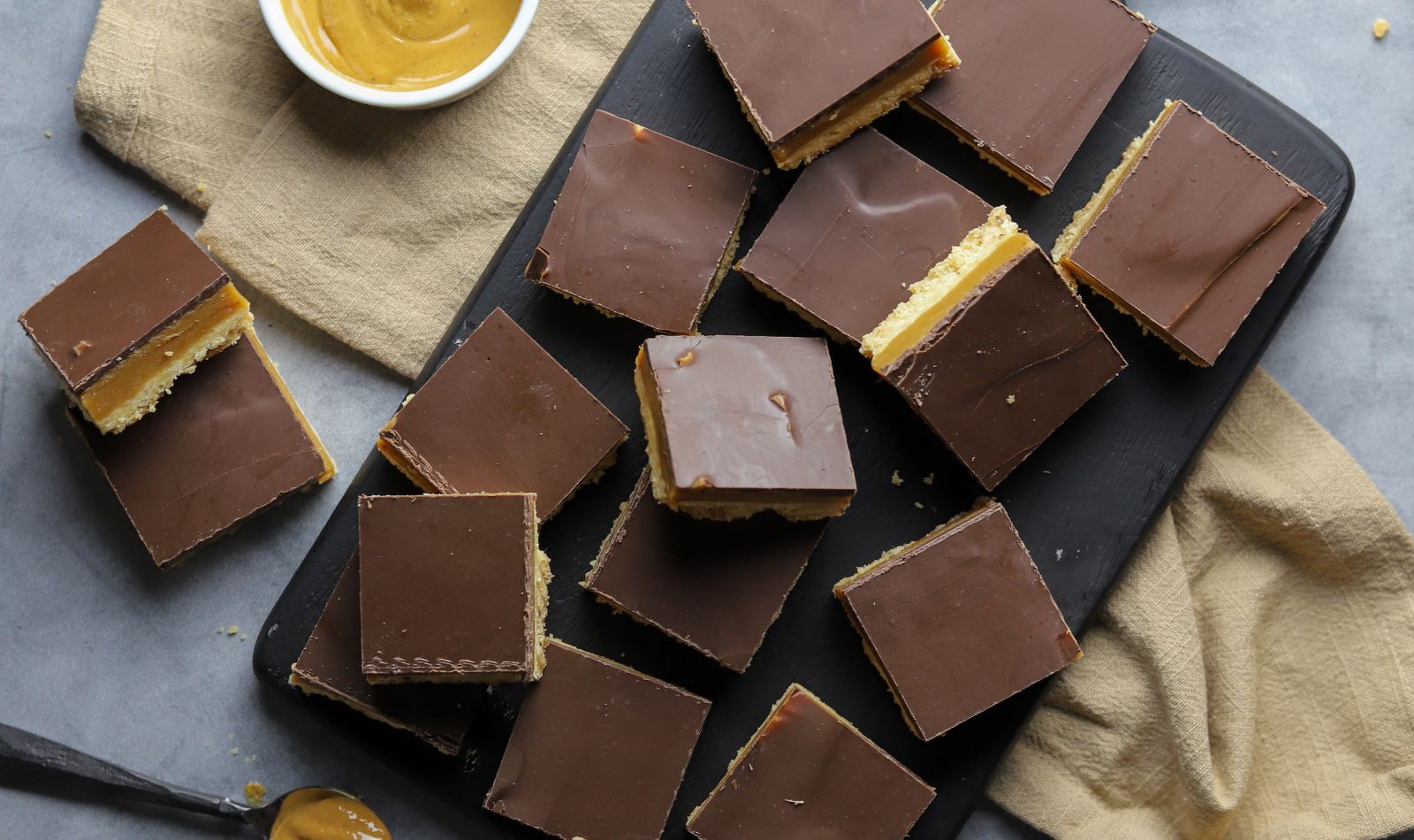 Peanut butter caramel shortbread cookies topped with milk chocolate, cut into squares, arranged on a black platter.