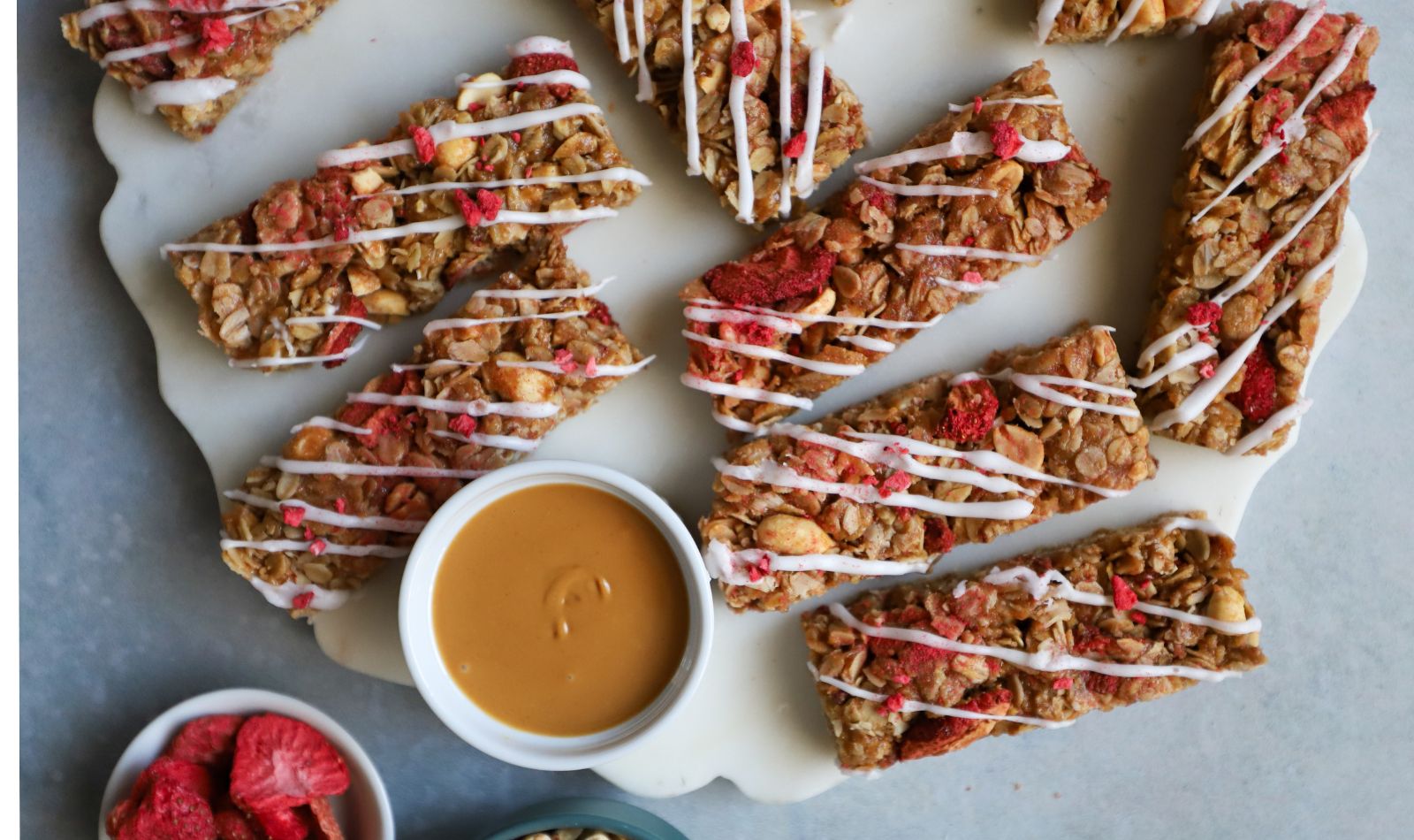 Crunchy peanut butter and strawberry granola bars with a sweet glaze, sprinkled with freeze-dried strawberries, arranged on a decorative marble platter with peanut butter and freeze-dried strawberries on the side.